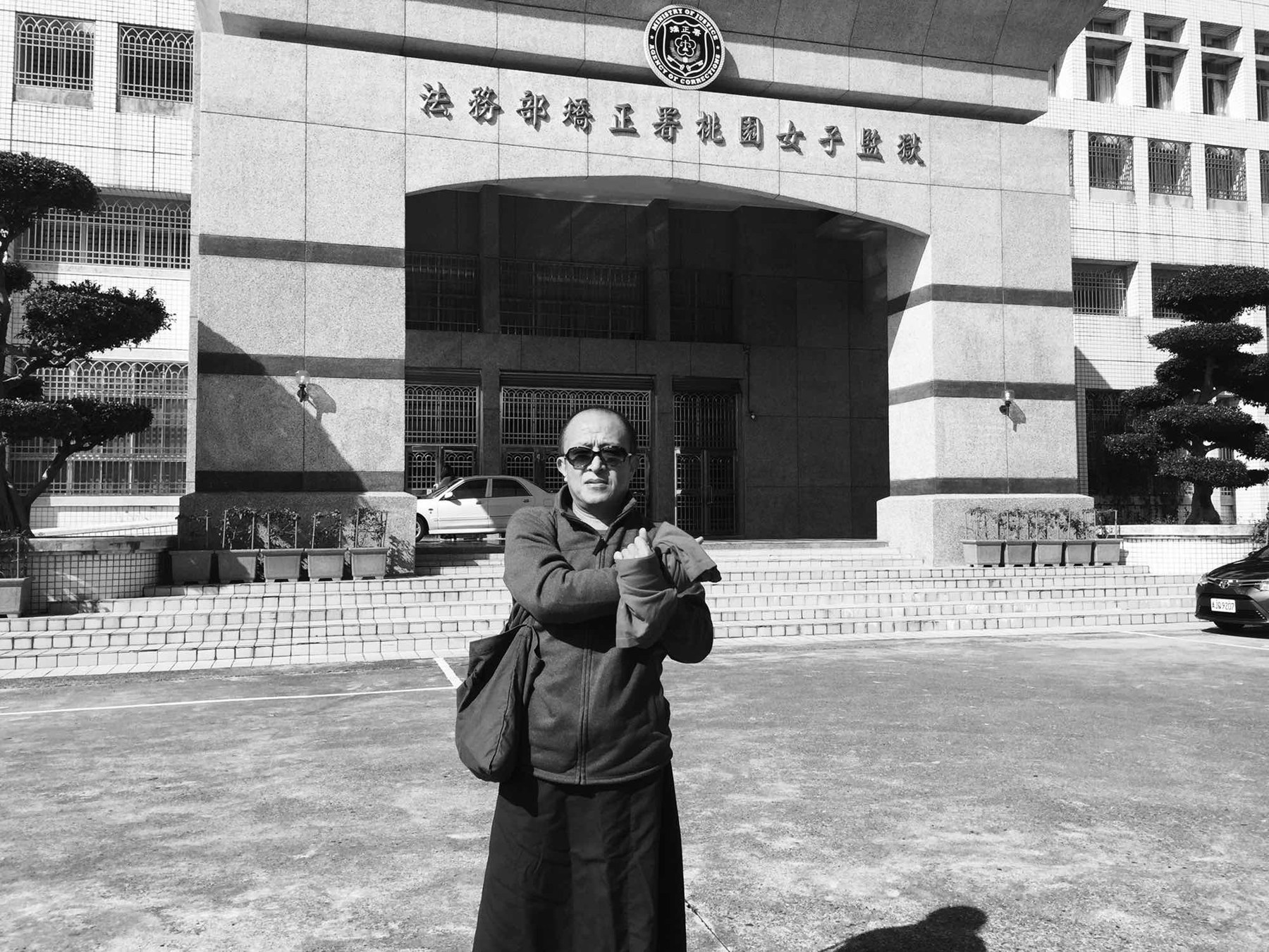 黑白照片中,仁波切戴墨镜,站在监狱门前将双手交叉伸出。
