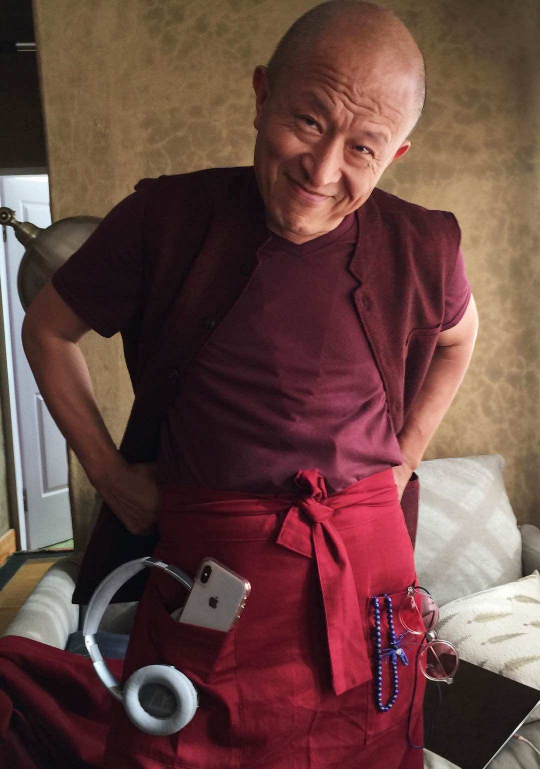 仁波切腰间系着装有手机与小器具的多口袋布件，双手放在腰侧。