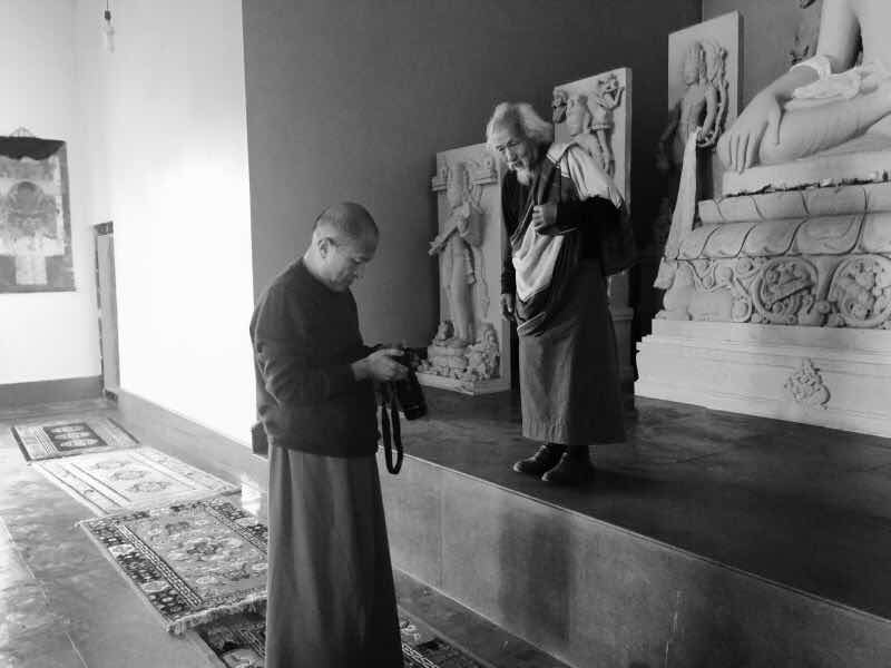 In a black-and-white photograph, two robed figures stand before Buddhist statues; one looks down at a camera.