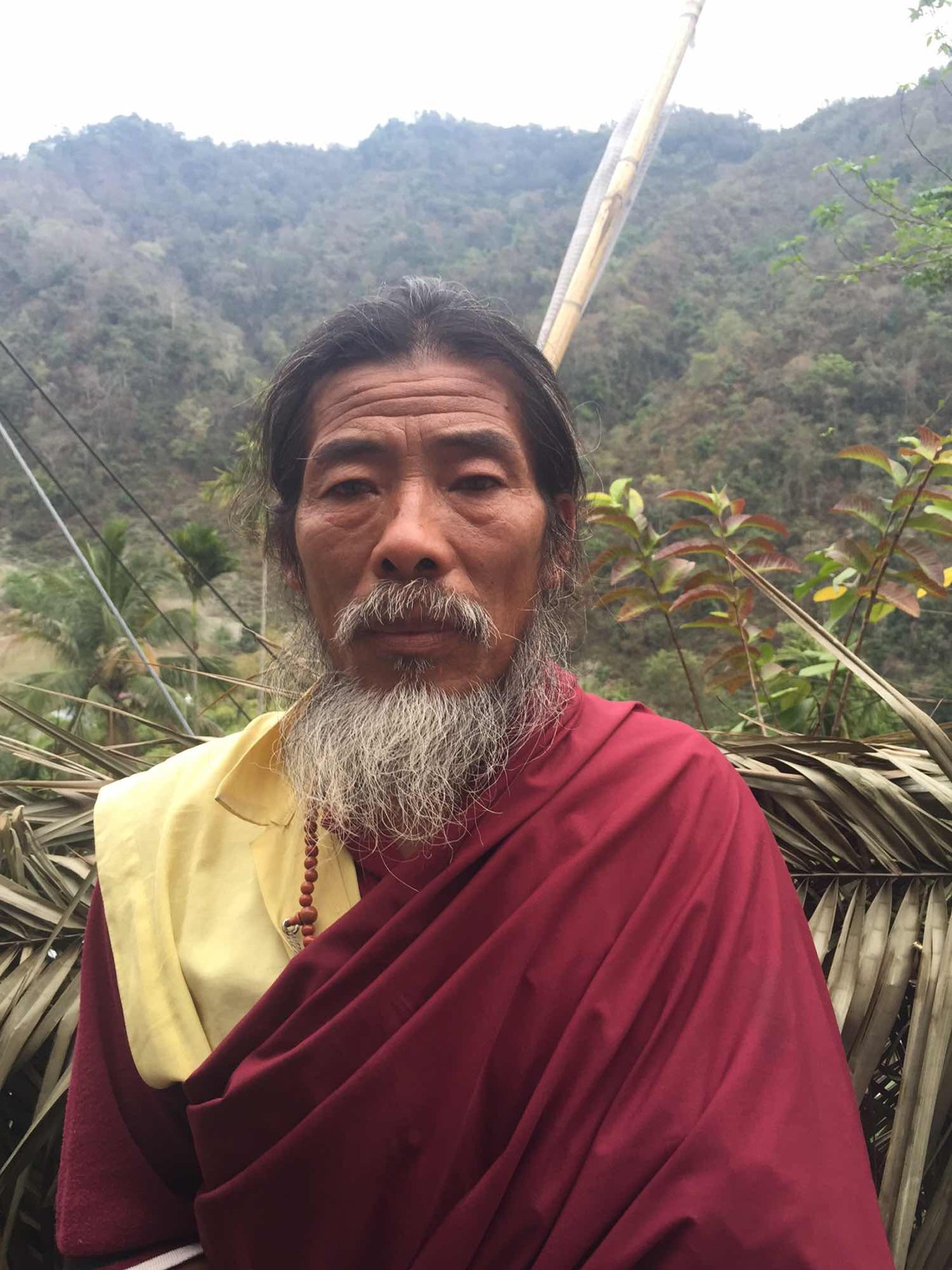 一位灰白胡须的人看向镜头，身后是山坡。